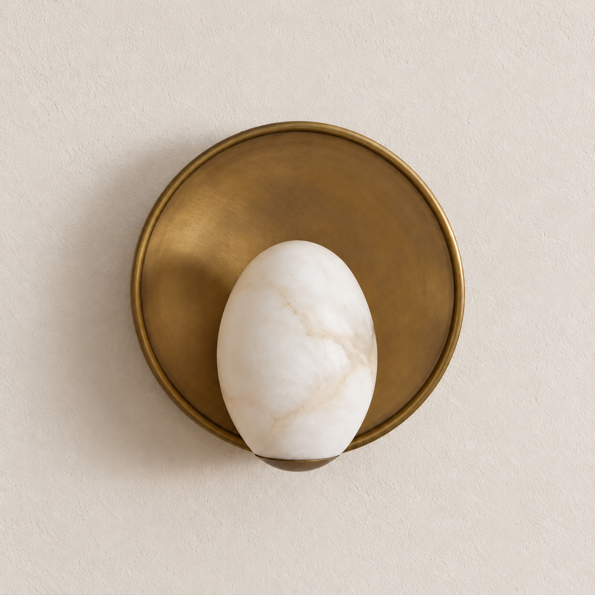 Unlit alabaster egg wall sconce against a round brushed-brass backplate on warm ivory.