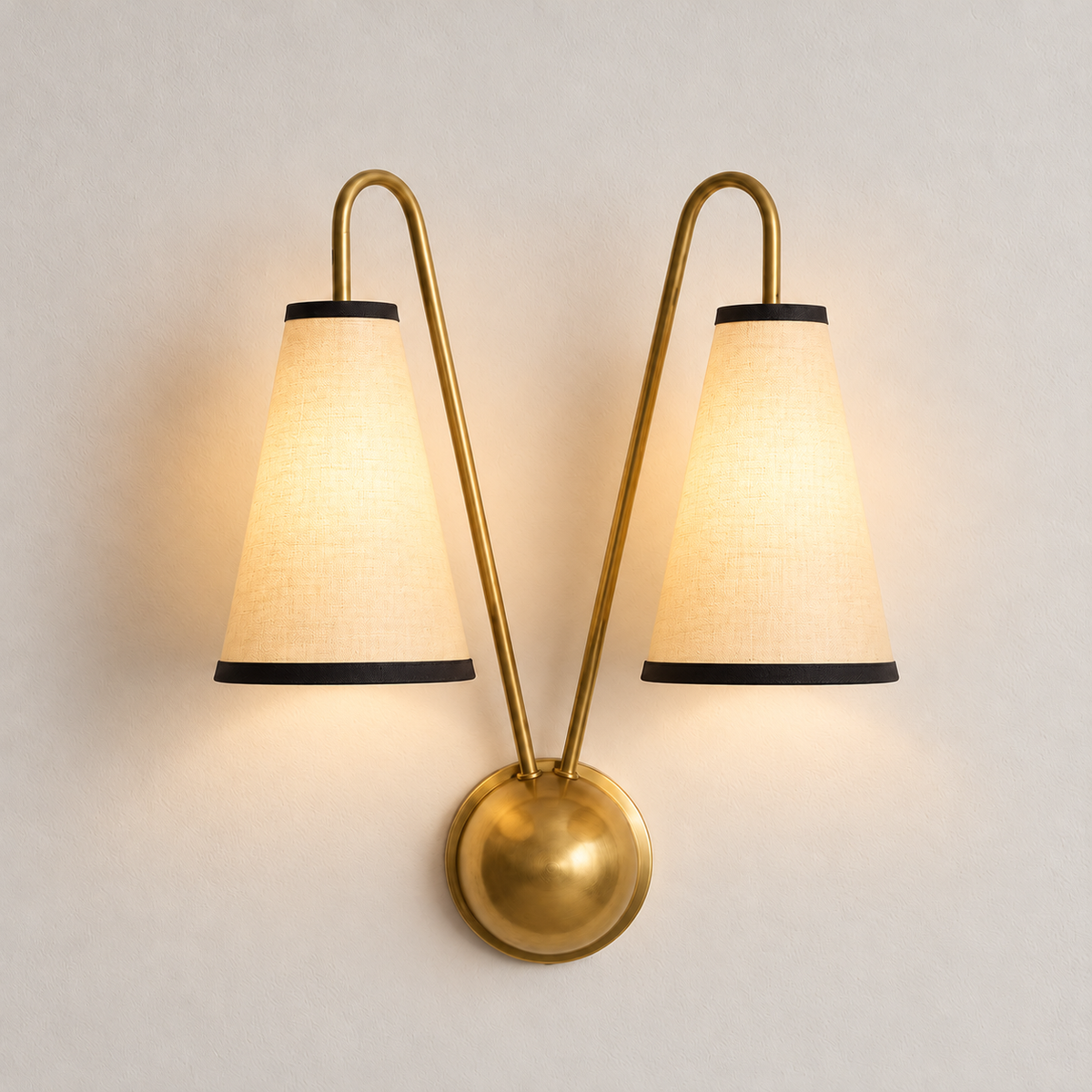 Brass twin-arm wall sconce with both cream fabric shades illuminated on warm ivory.