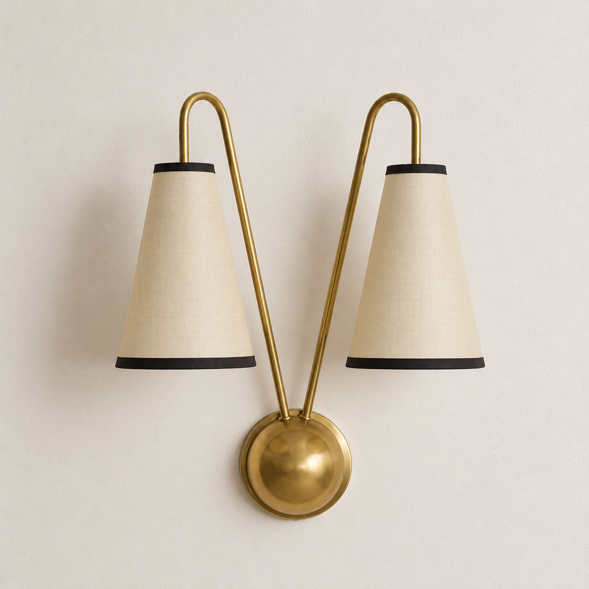 Unlit brass twin-arm wall sconce with two cream fabric shades and dark trim on warm ivory.