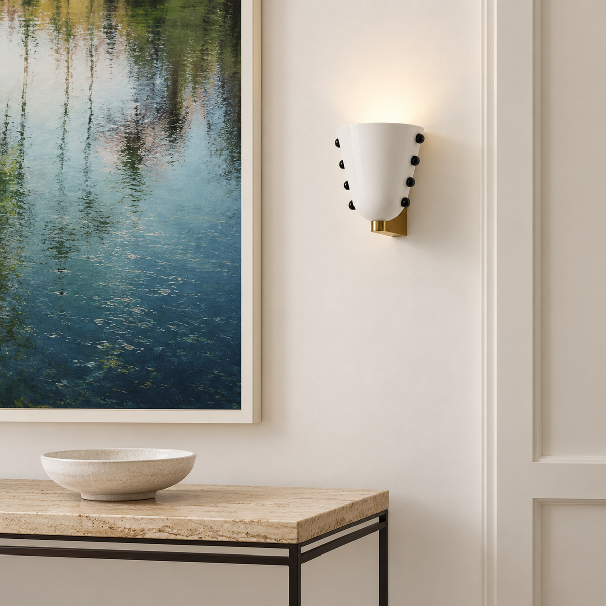 White glass wall sconce with black studs and gold brass mount, installed beside artwork above a console