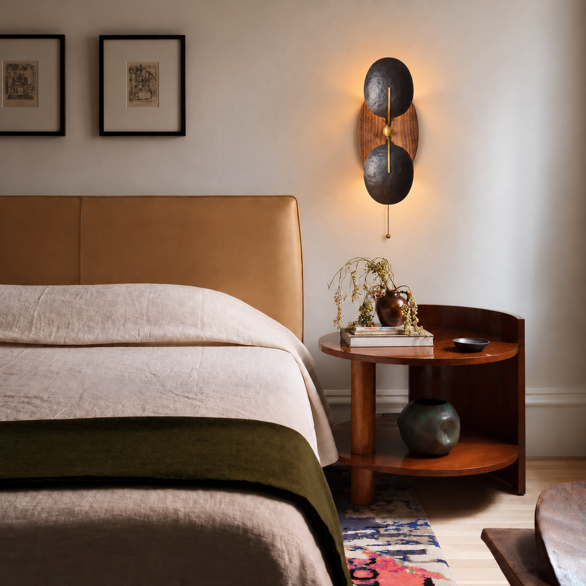 Front view of an illuminated walnut and iron wall sconce mounted flat above a round walnut bedside table.