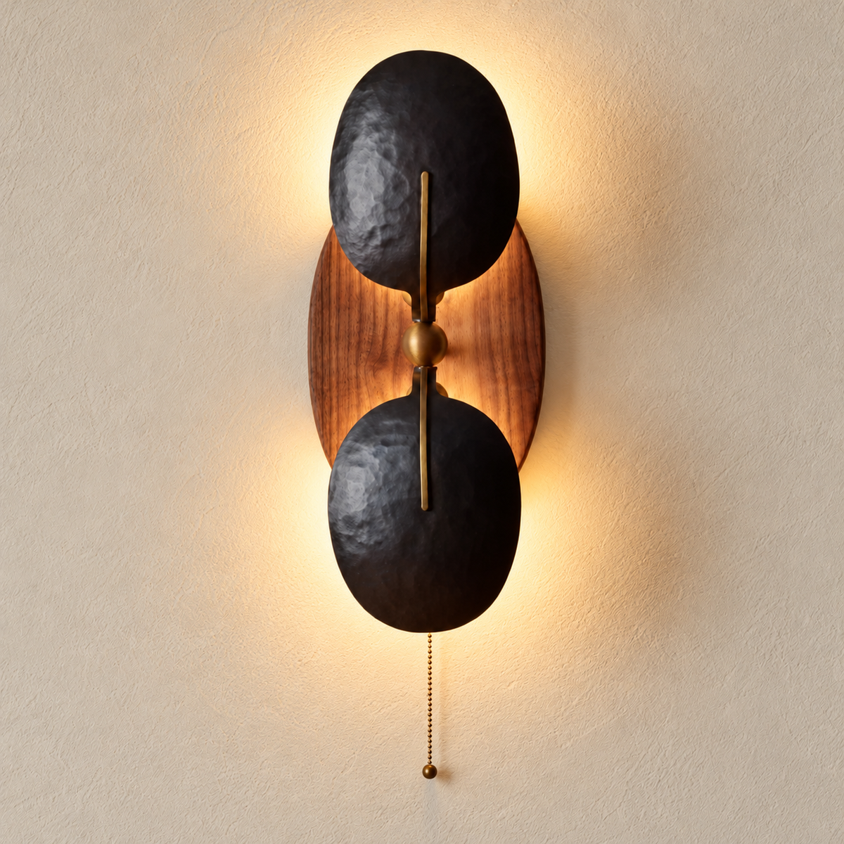 Walnut and hammered iron wall sconce illuminated with warm light behind its two opaque shades, with the pull chain visible.