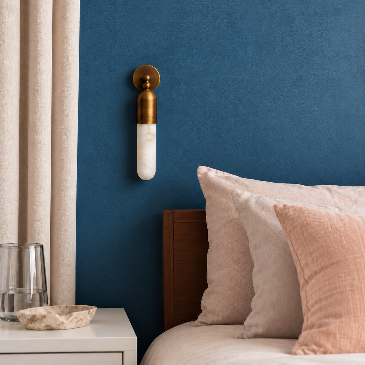 Brass and alabaster tube sconce mounted beside blush bedding on a blue bedroom wall.