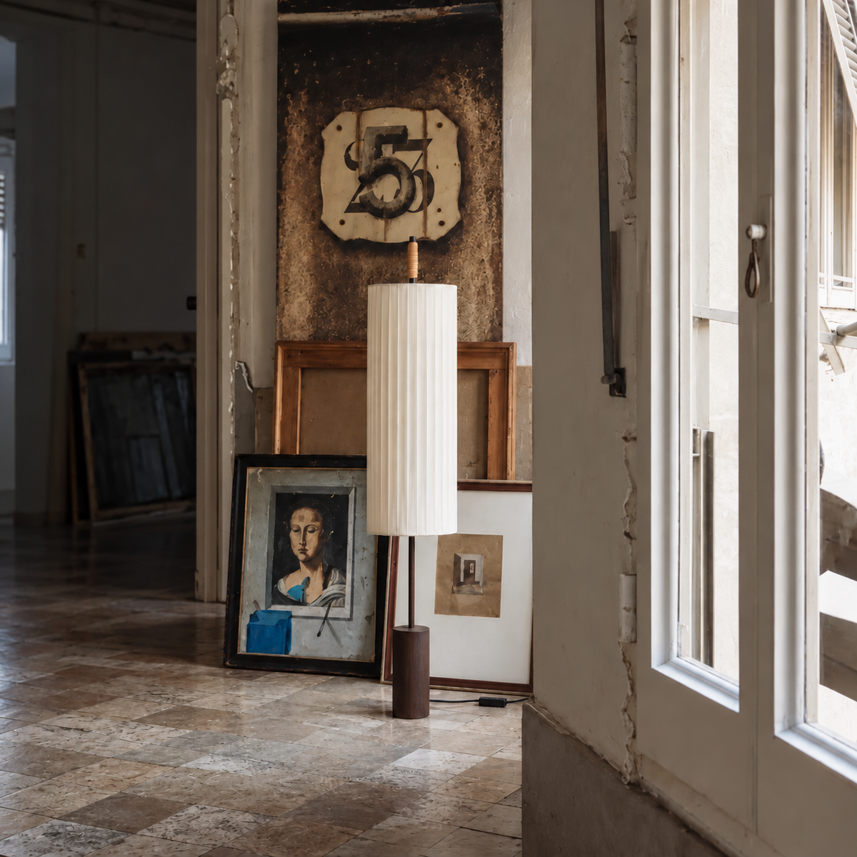 CLHBV0255 floor lamp standing among leaning framed artworks beside a tall studio window.