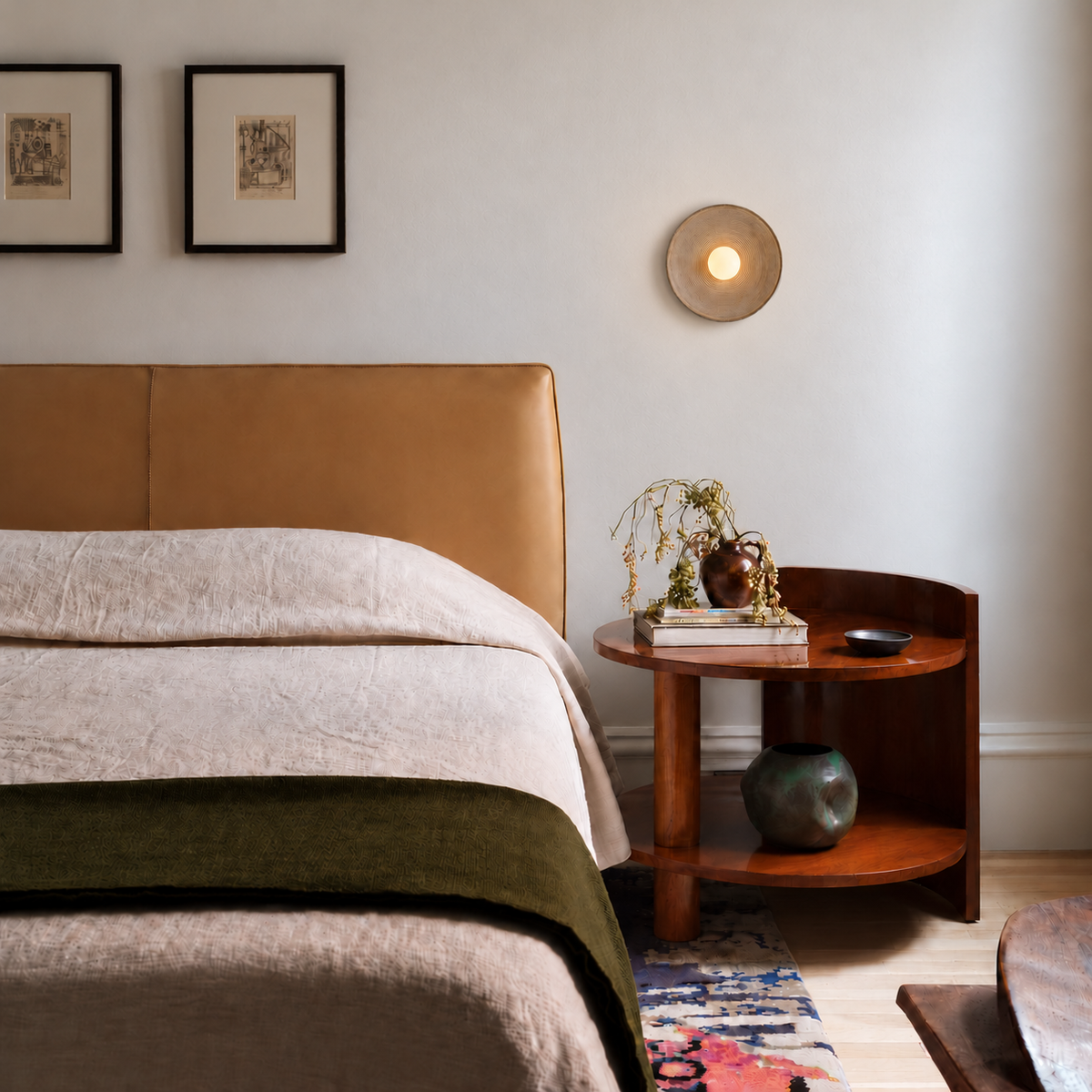 Taupe ceramic disc wall lamp mounted above a walnut bedside table in a bedroom lit by natural daylight.