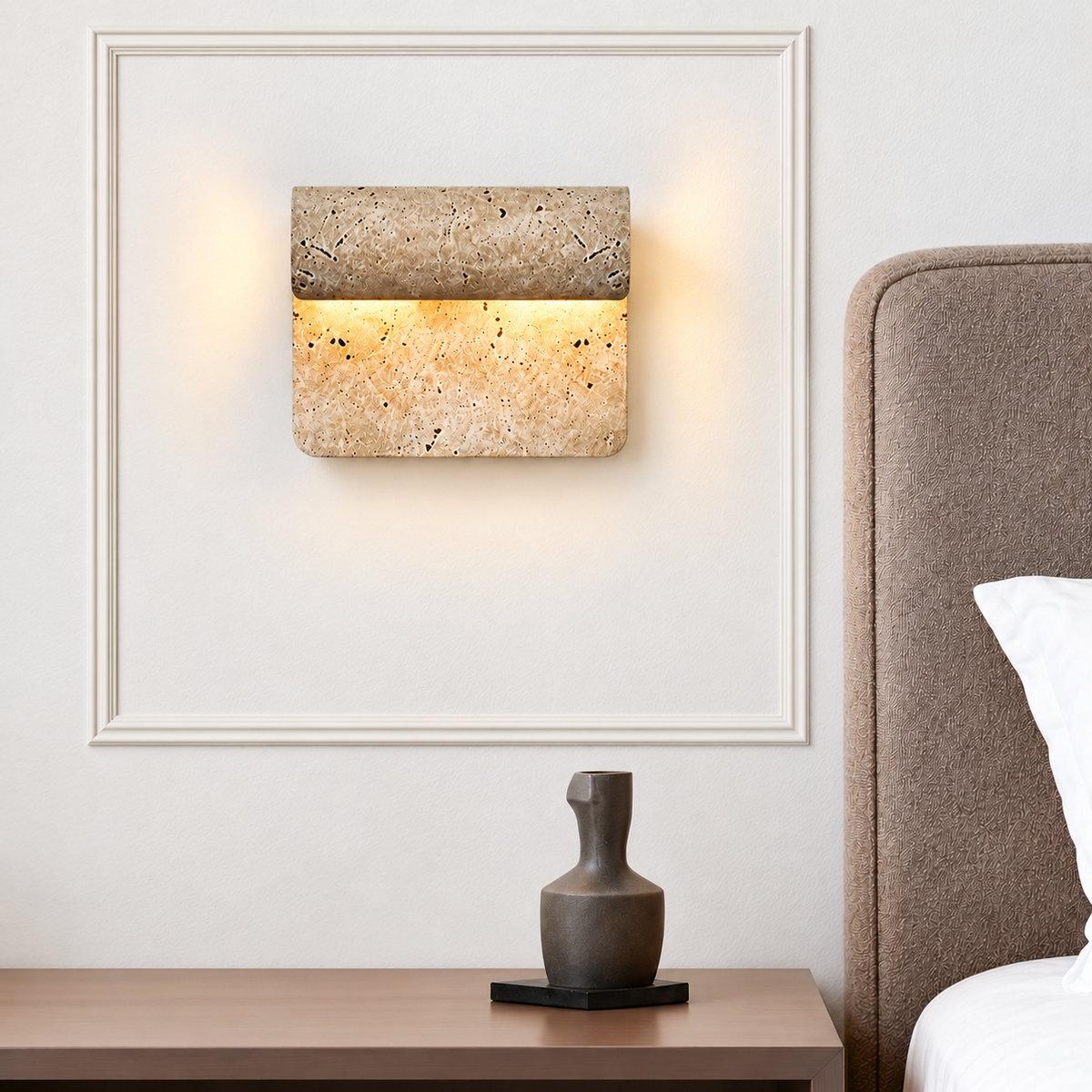 Curved travertine wall sconce beside an upholstered bed, framed by ivory wall mouldings.